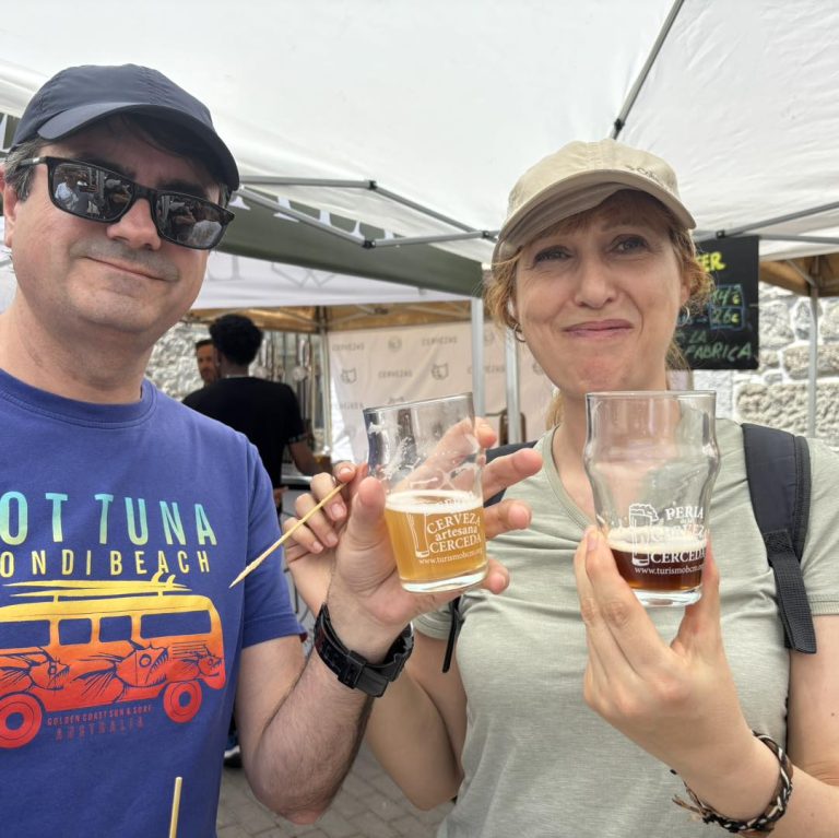Maestro cervecero explicando la elaboración de cerveza artesana a entusiastas visitantes en un puesto de la feria, con equipos cerveceros en el fondo.